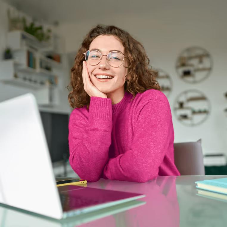 Eine junge Frau mit Brille sitzt lachend an einem Glastisch vor ihrem Laptop