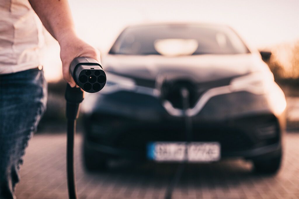 Eine Hand hält einen Ladestecker für ein Elektroauto vor dem Fahrzeug