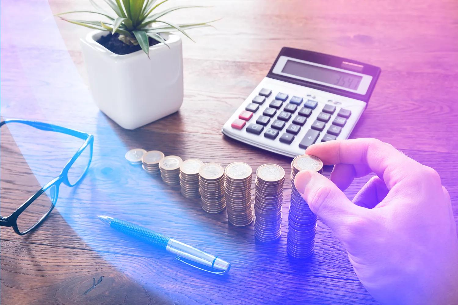 A hand stacks coins next to a calculator, a pair of glasses and a pen on a wooden table