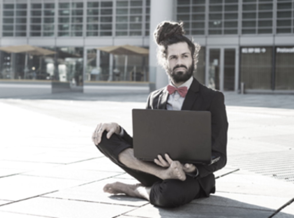 Ein Mann im Anzug mit Fliege sitzt barfuß im Schneidersitz auf dem Boden und arbeitet an einem Laptop