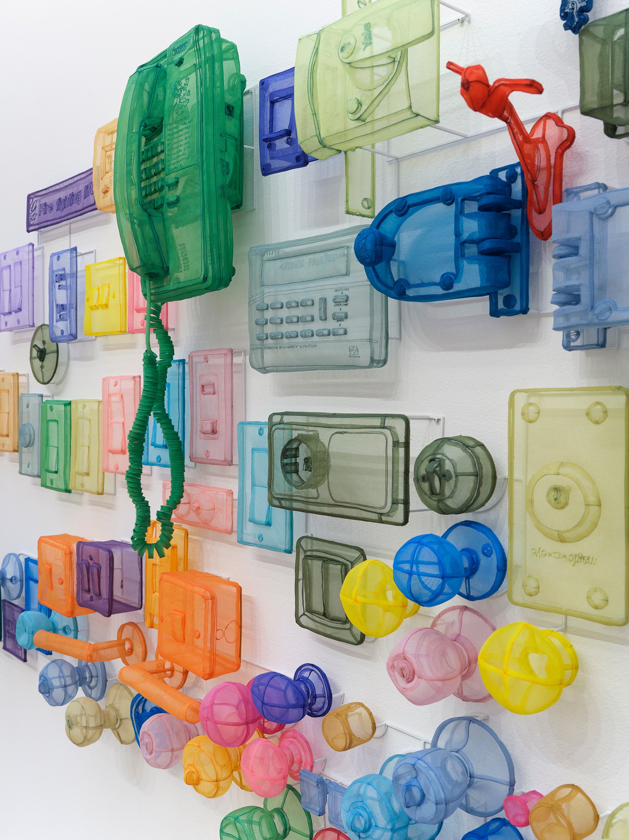 A wall-mounted installation of translucent colored plastic objects — vintage telephone handsets, electrical outlet covers, and spherical forms — arranged against a white wall.
