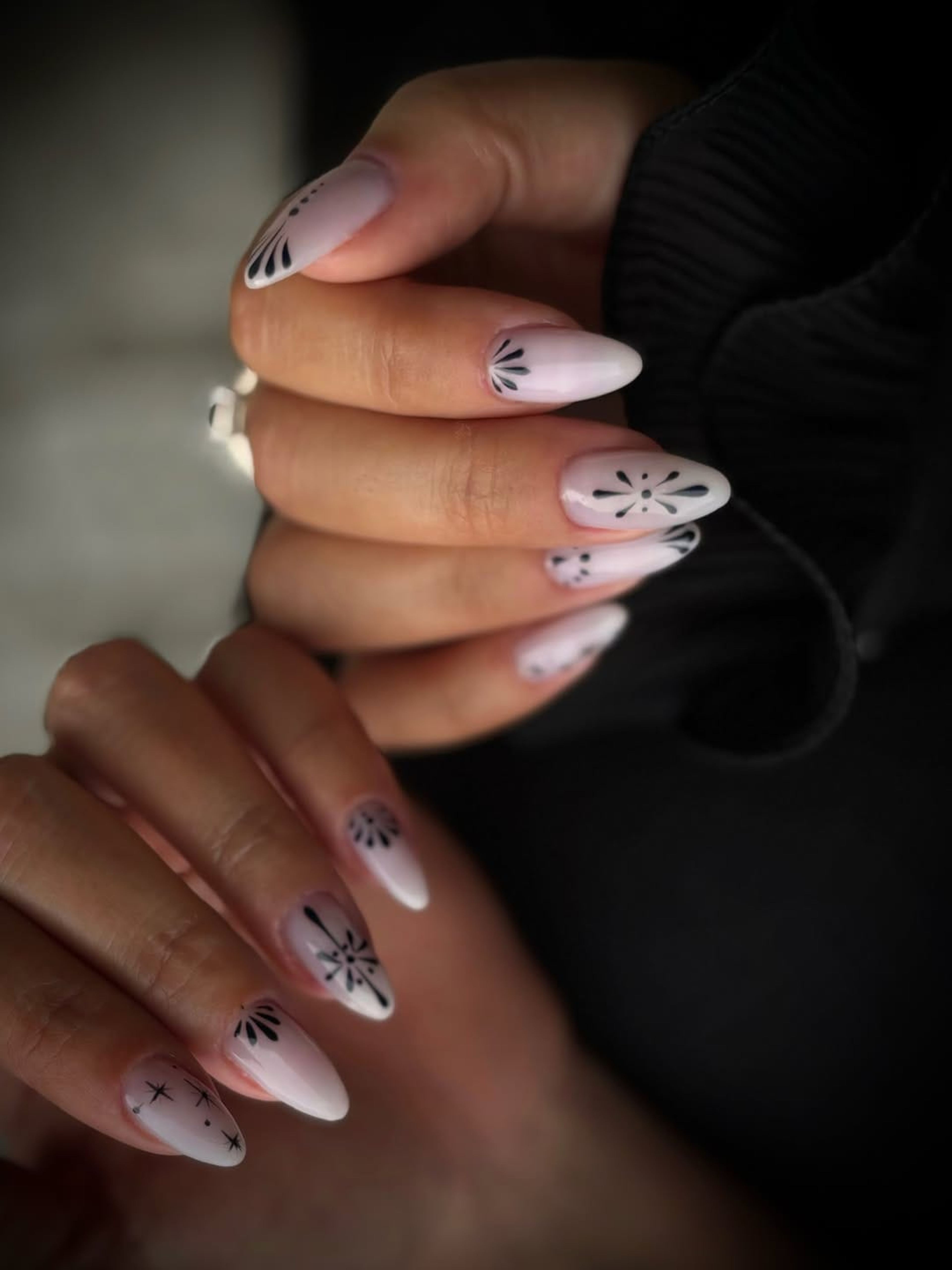 Ongles amande nude ornés d’un motif noir fin, sur fond sombre.