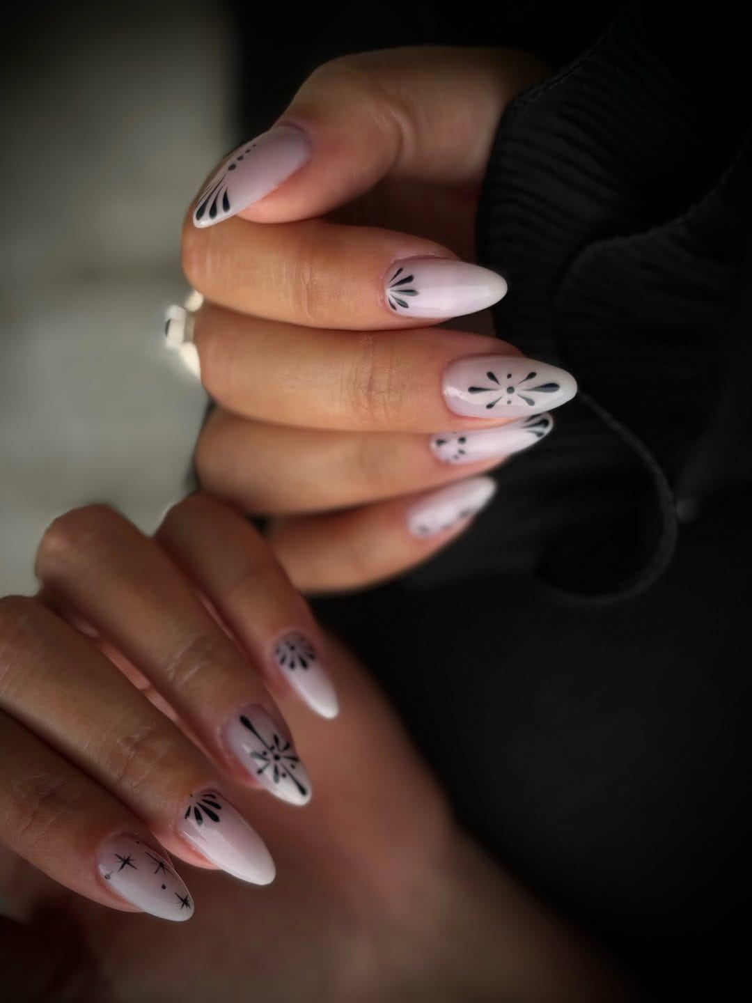 Ongles amande nude ornés d’un motif noir fin, sur fond sombre.