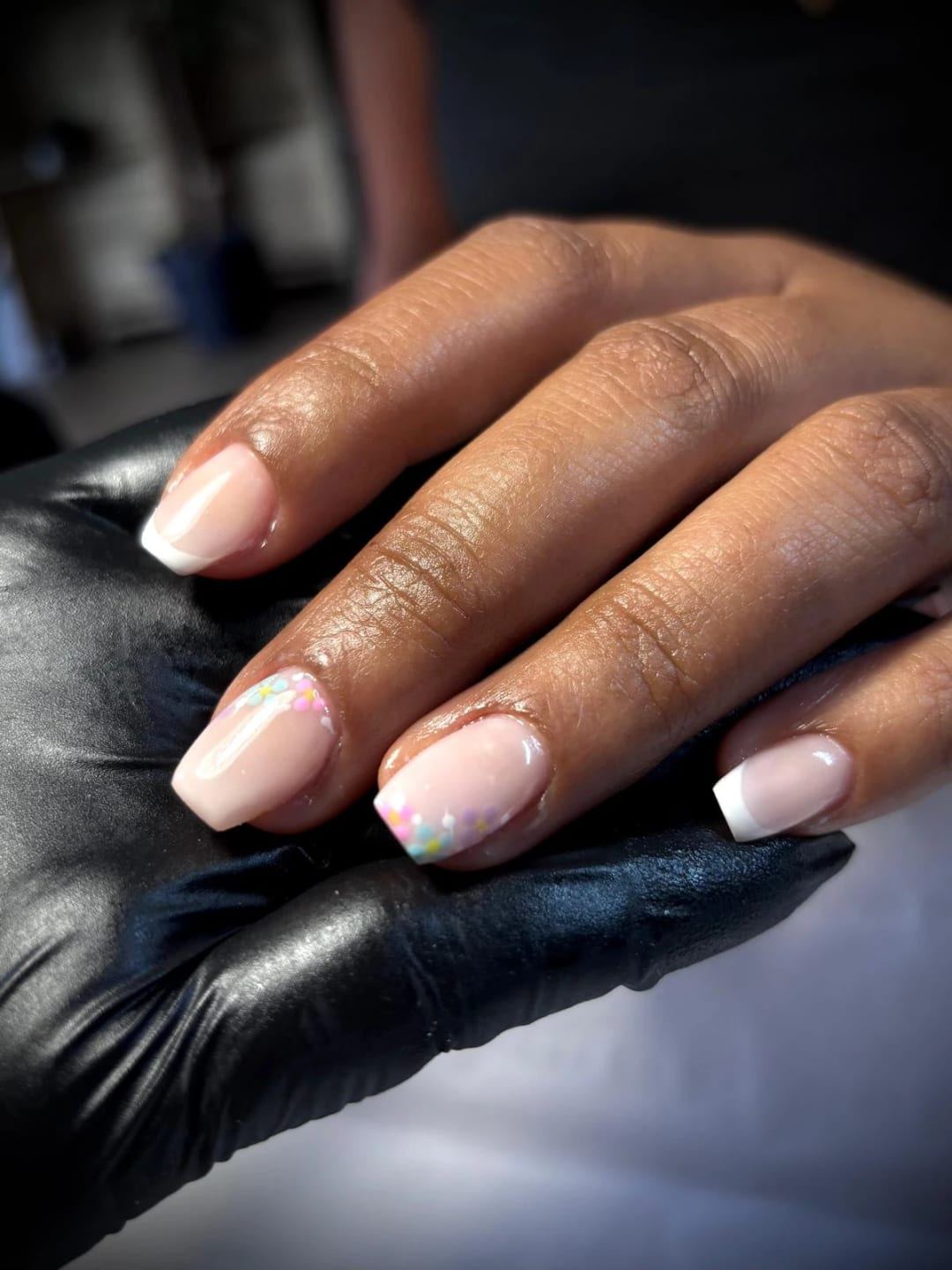 Main aux ongles en french blanche, fleurs pastel peintes en bord d’ongle, posée sur une main gantée de noir.