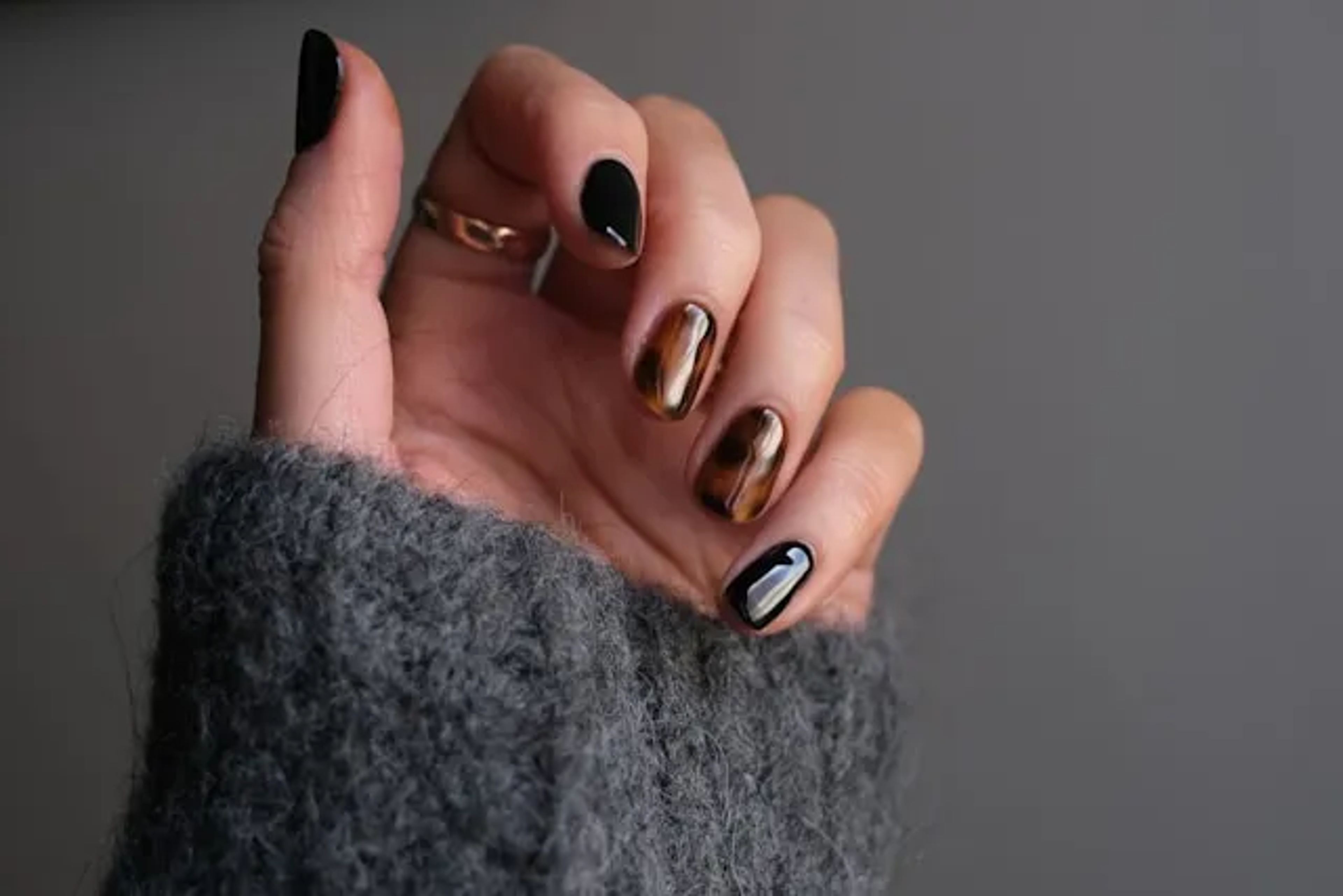 Main aux ongles noirs et cuivrés à effet miroir, sortant d’une manche en laine grise