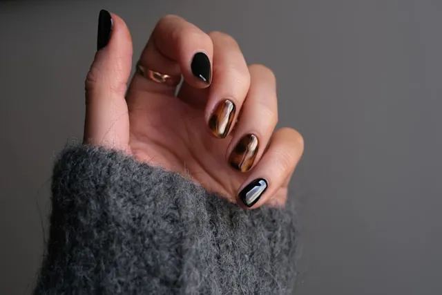 Main aux ongles noirs et cuivrés à effet miroir, sortant d’une manche en laine grise