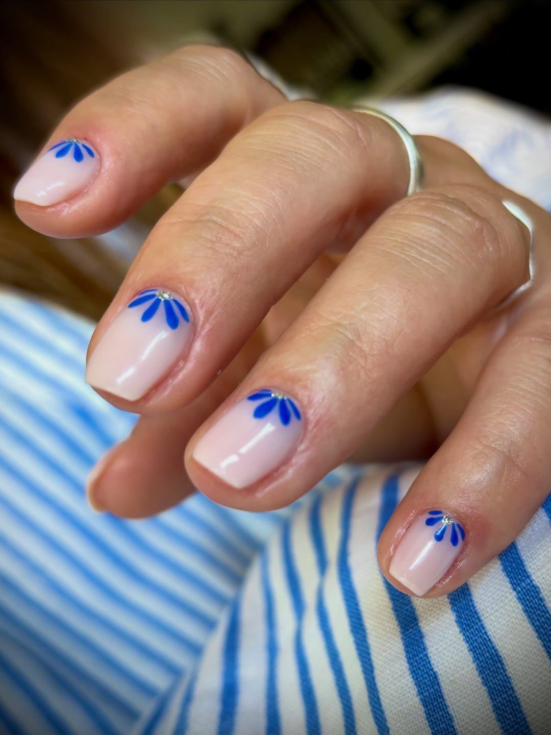 Ongles courts nude ornés de pétales bleus, sur une chemise rayée.