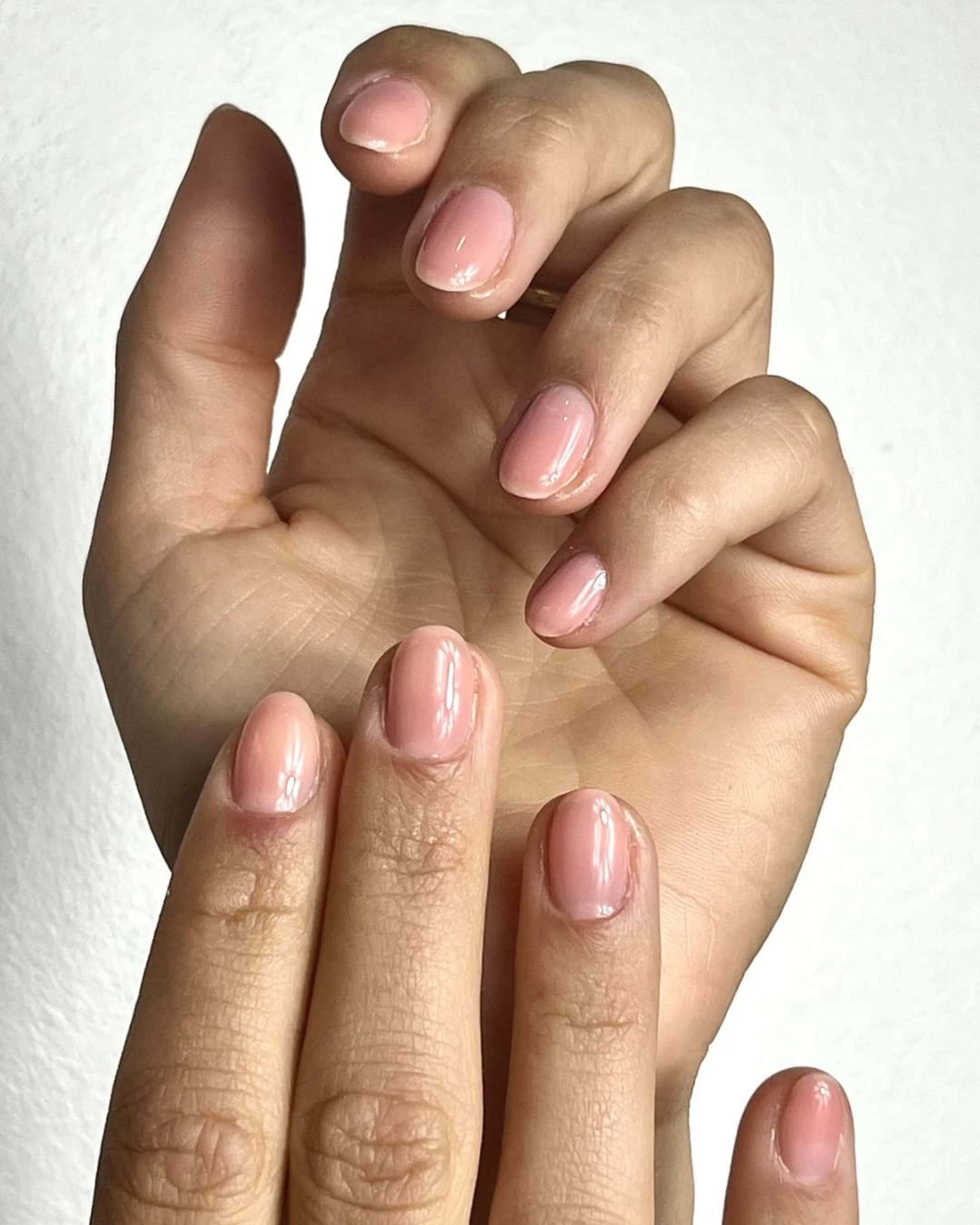 Deux mains posées l’une sur l’autre, ongles courts en gel nude rosé, sur fond blanc.