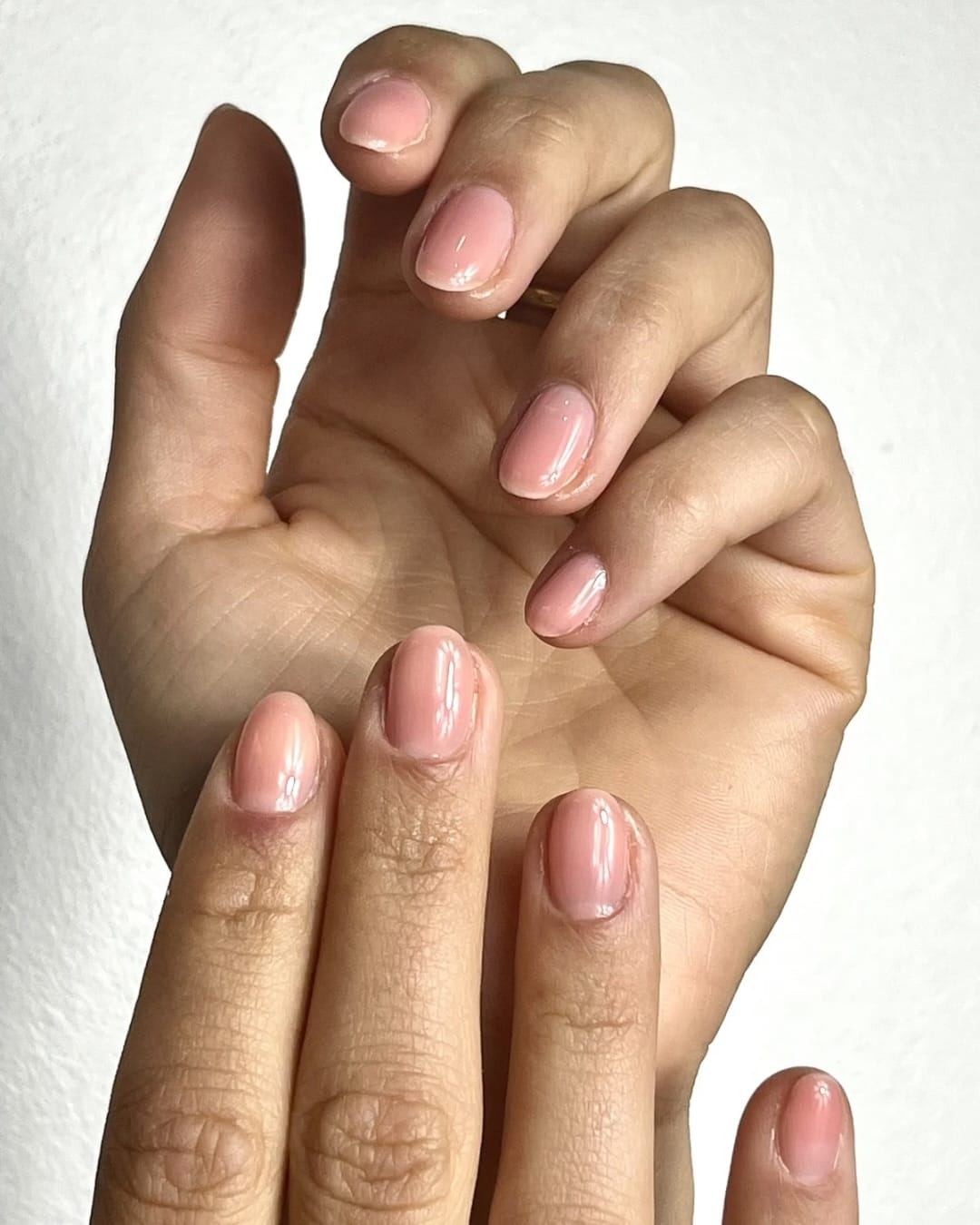 Deux mains posées l’une sur l’autre, ongles courts en gel nude rosé, sur fond blanc.