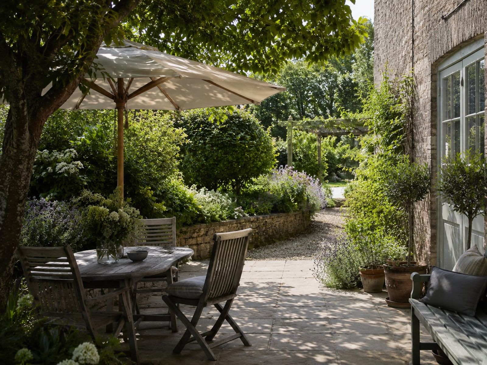 A shaded stone terrace beneath a parasol and pergola, planting softening the heat beside the house.