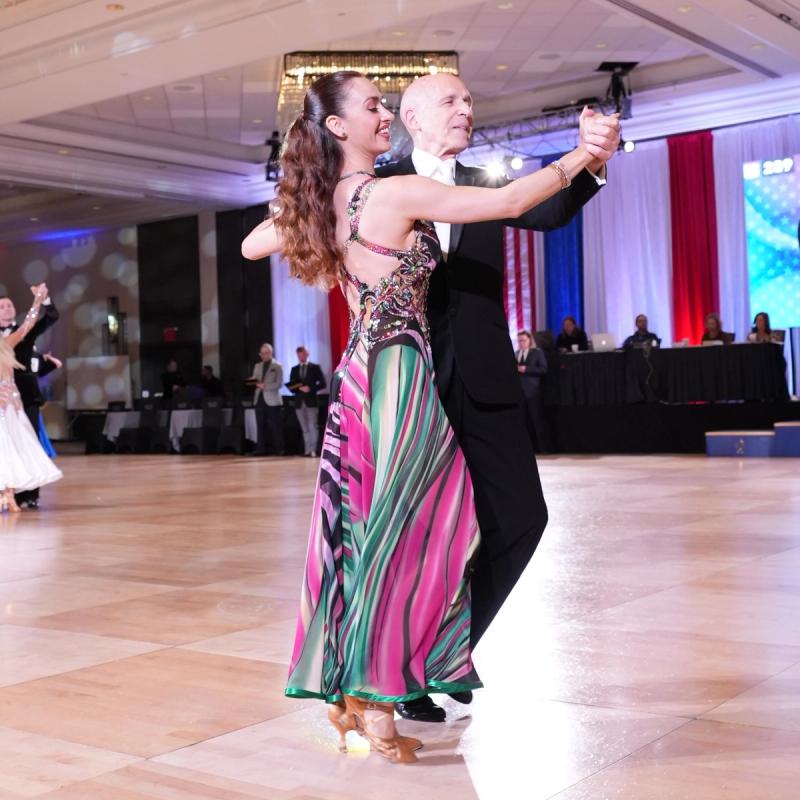 A couple dancing a smooth ballroom routine, the follower in a flowing pink and green gown, the leader in a tailsuit.