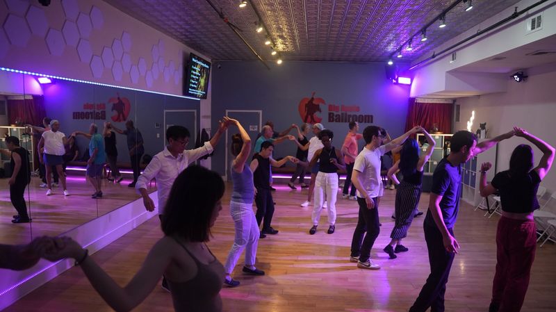 A West Coast Swing group class at Big Apple Ballroom, couples practising in loose rows under purple lighting with the studio’s logo on the wall behind.