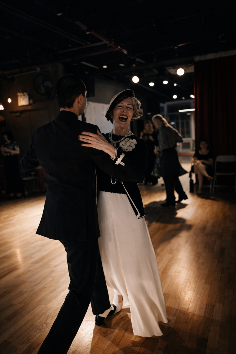 A couple dancing Foxtrot in closed position at Big Apple Ballroom, the follower in a long cream gown, both laughing.