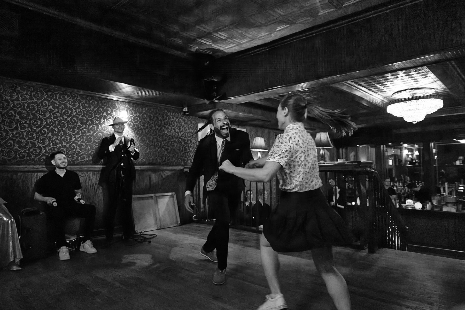 A swing couple mid-step at the NY Nightlife Showcase — her ponytail flying, the band clapping along, in black and white.