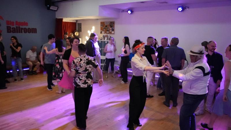 Dancers swinging out on the Big Apple Ballroom floor, one in a red headscarf and another in a white fedora, under purple lights.