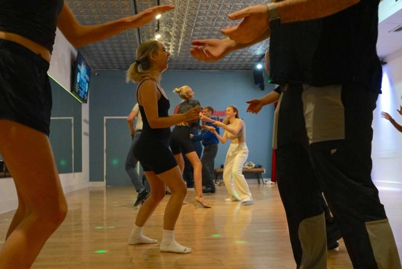 Dancers in a Bachata class at Big Apple Ballroom, a student laughing mid-step in the centre while others practise in pairs across the wooden floor.