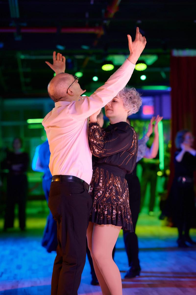 A couple dancing with both arms raised, lit in green and teal, at a Big Apple Ballroom social.
