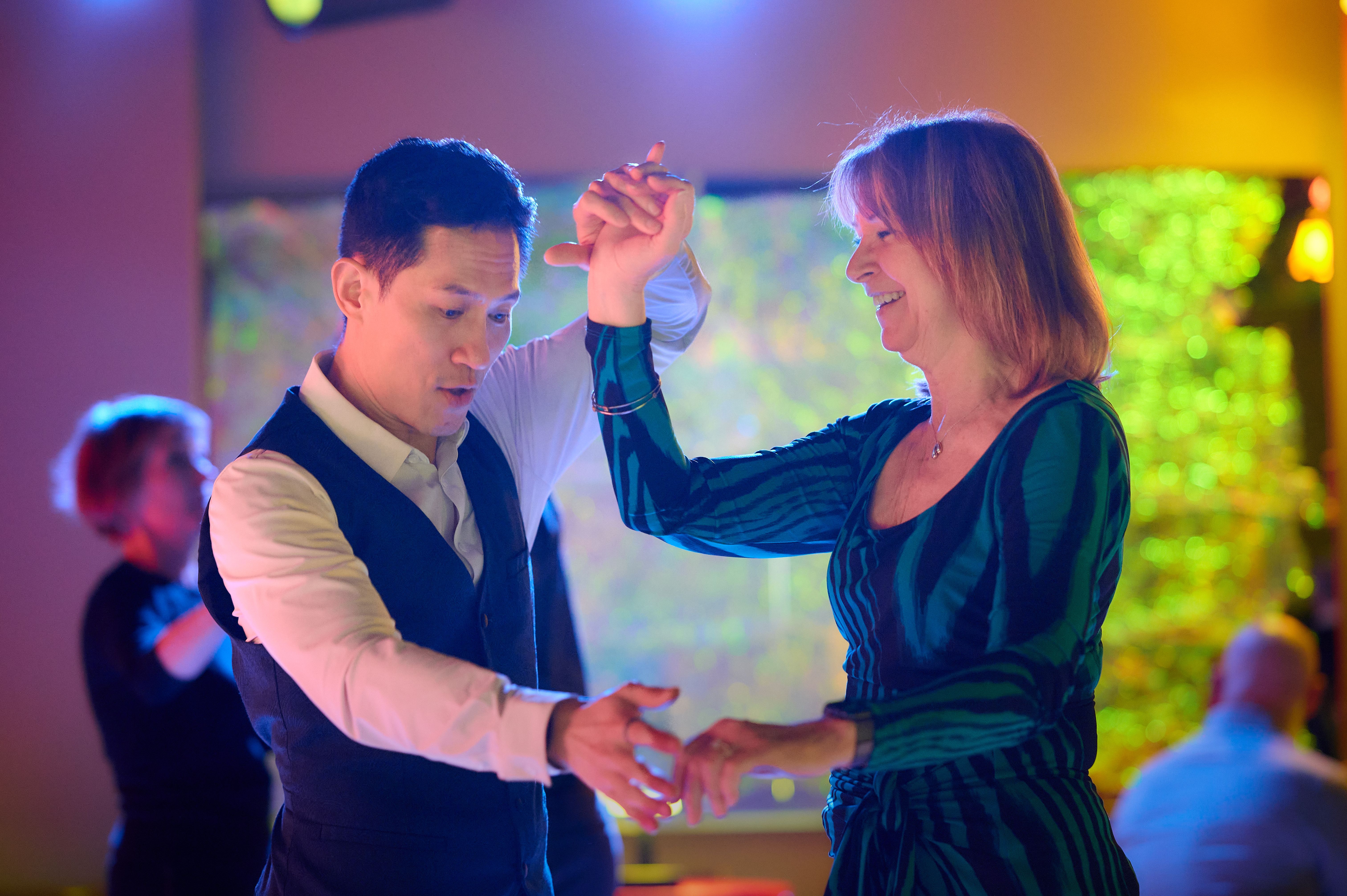 A couple mid-dance at a Big Apple Ballroom social, smiling at each other under colorful party lighting.