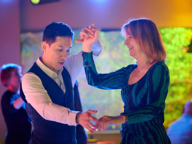 A couple mid-dance at a Big Apple Ballroom social, smiling at each other under colorful party lighting.