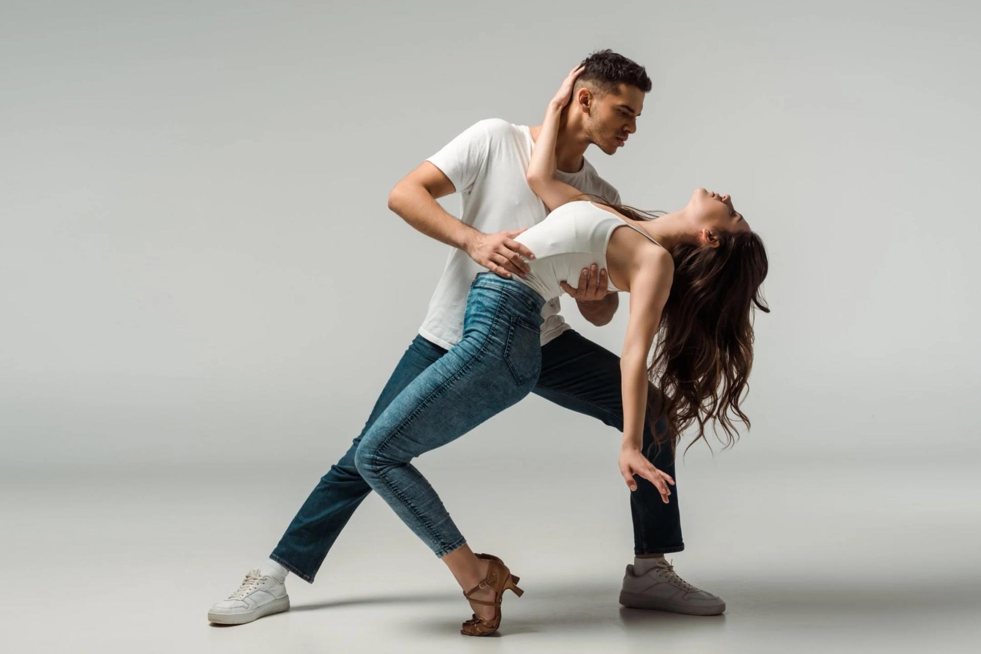 A couple in casual dress in a deep rumba dip — she arches back over his arm, studio-lit against a plain backdrop.
