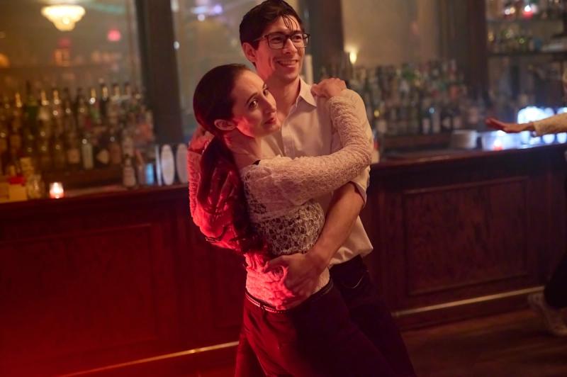A couple dancing in a close hold and smiling, in front of a warmly lit speakeasy bar.
