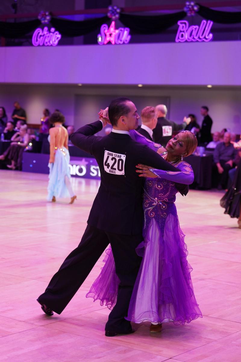 A couple competing in ballroom, the leader in a tailsuit and the follower in a flowing purple ballgown.