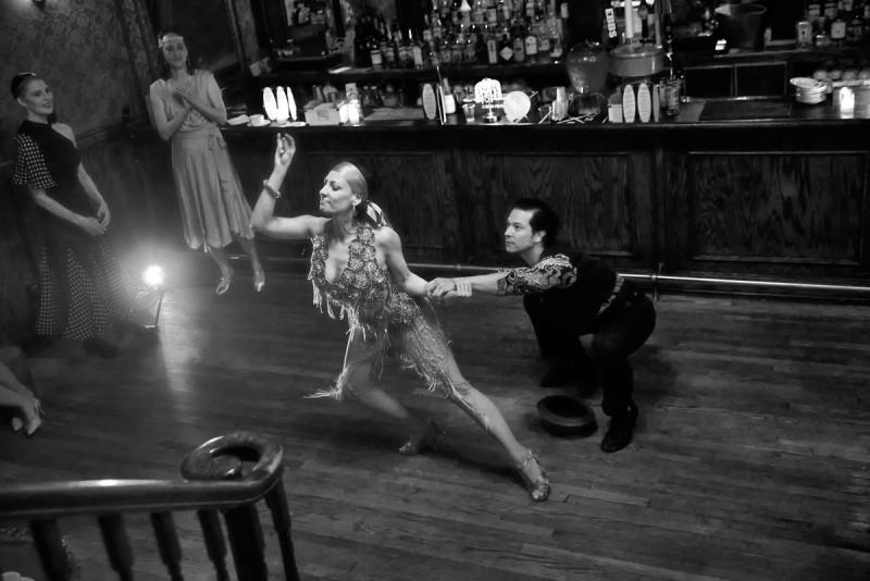 Black and white photograph of two dancers in a deep extended line, the follower reaching forward in a fringed dress, at a speakeasy bar.