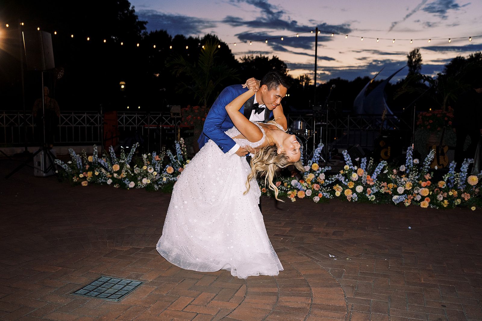 A groom in a blue suit dips his bride in her white gown during their first dance at dusk, flowers and string lights behind them.
