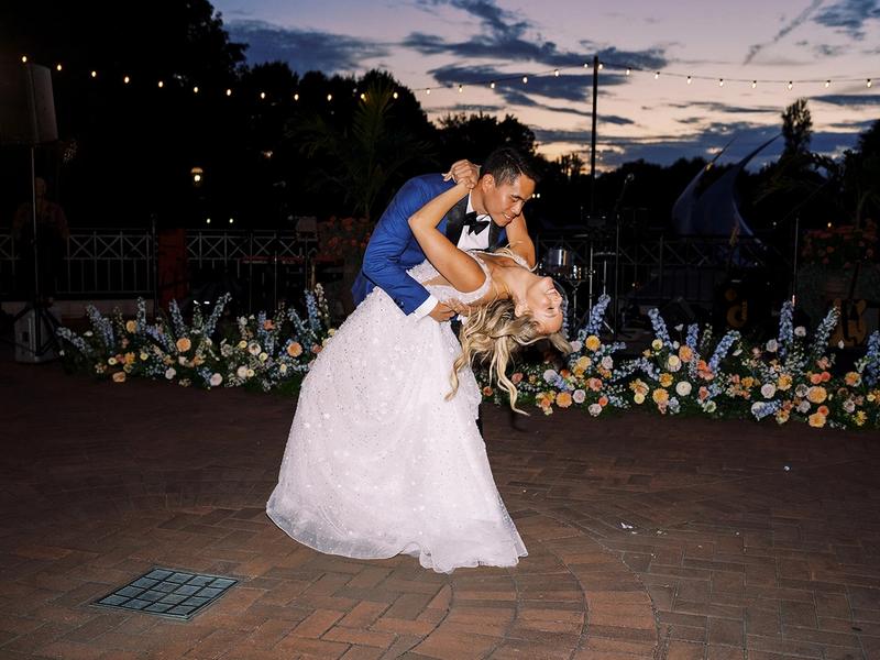 A groom in a blue suit dips his bride in her white gown during their first dance at dusk, flowers and string lights behind them.