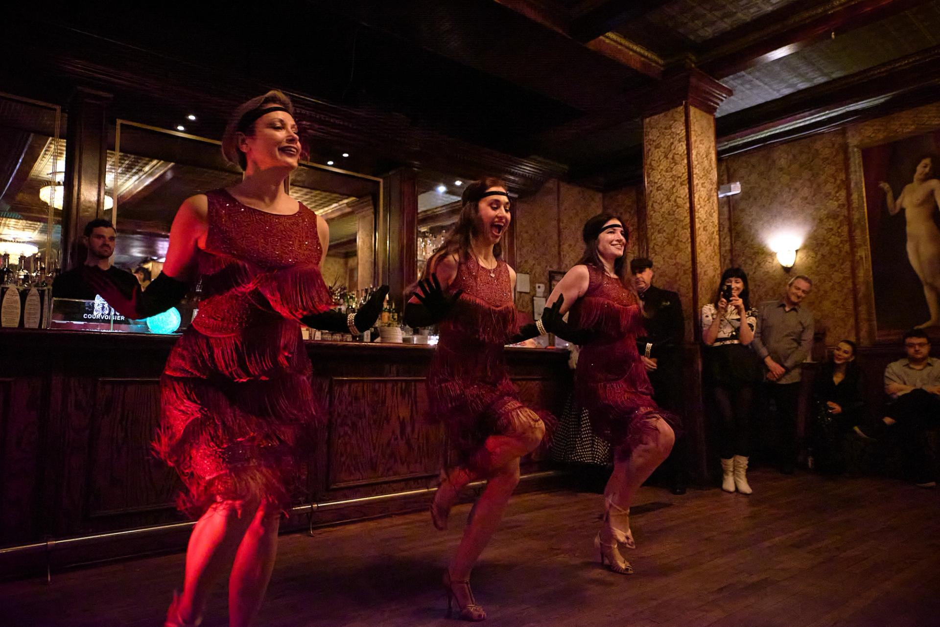 Three dancers in fringe flapper dresses and headbands mid-Charleston kick at the NY Nightlife Showcase bar.
