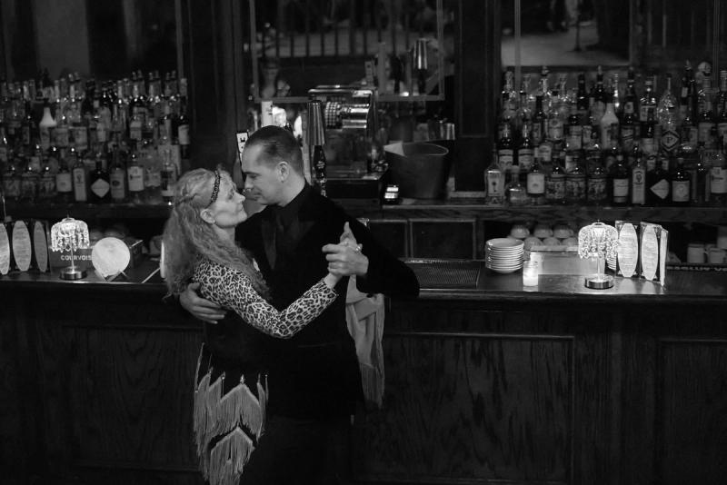 Black and white photograph of a couple in a close tango embrace, cheek to cheek, in front of a bar.