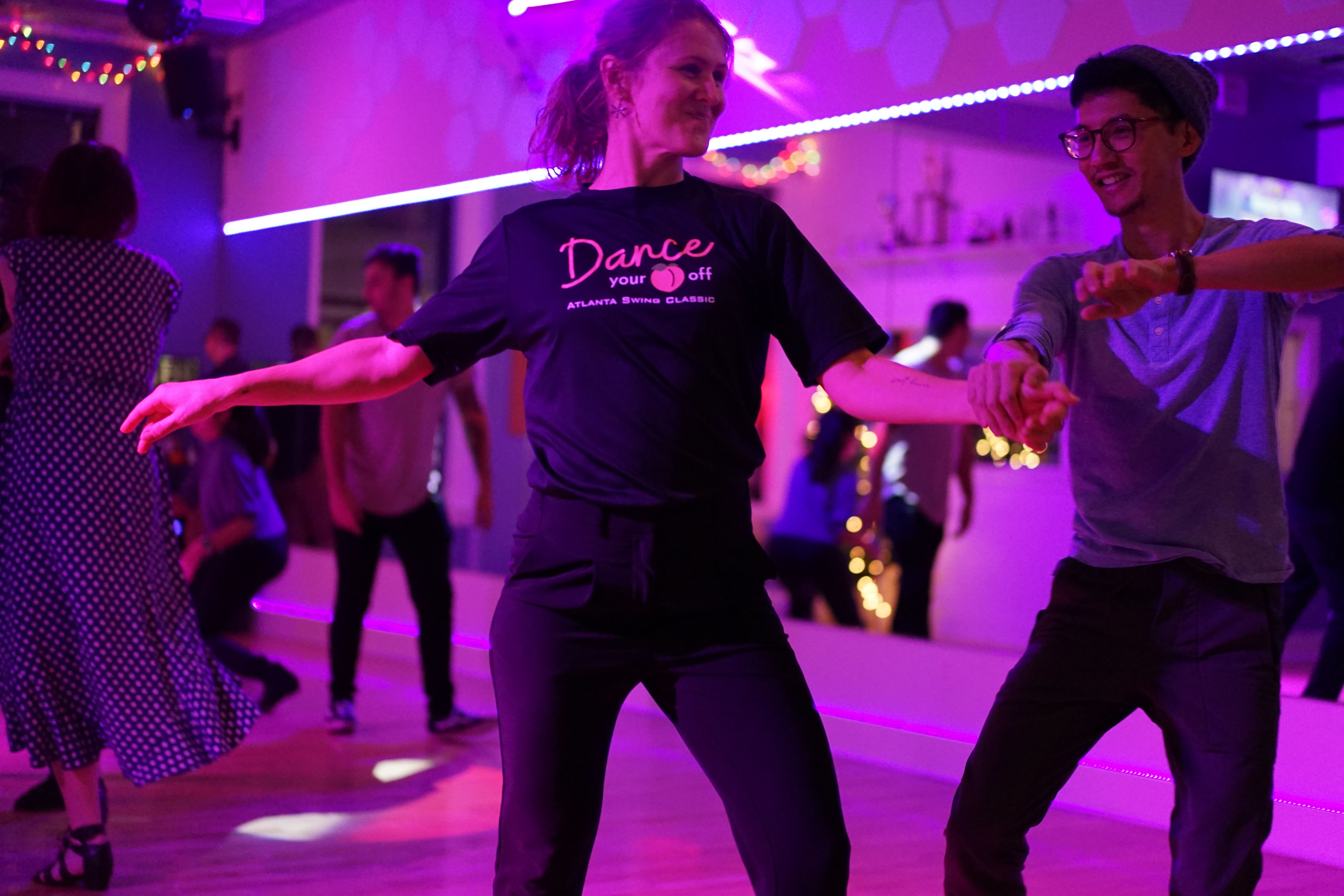 Two swing dancers mid-move at a studio party, arms stretched and grinning, colorful lights across the floor.