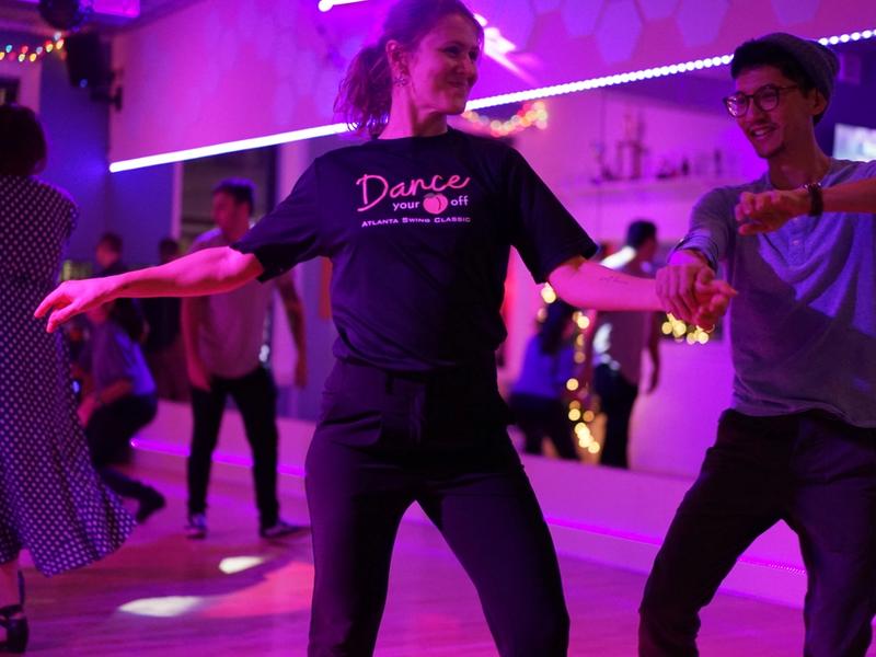 Two swing dancers mid-move at a studio party, arms stretched and grinning, colorful lights across the floor.