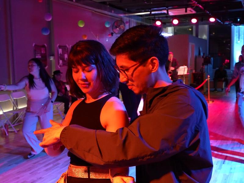 Two dancers in a close hold at a Big Apple Ballroom social night, blue and red light across the floor.