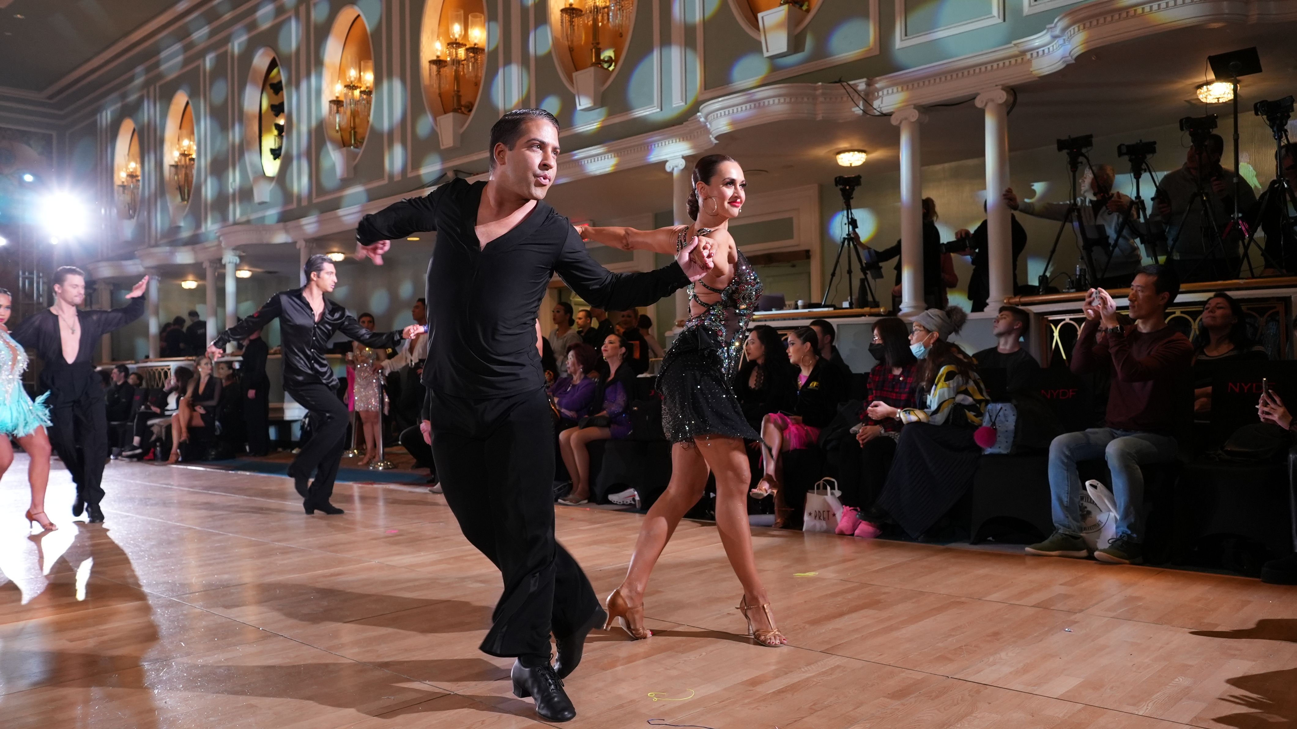 A Latin couple mid-heat on the New York Dance Festival competition floor, spectators and photographers watching from ringside.