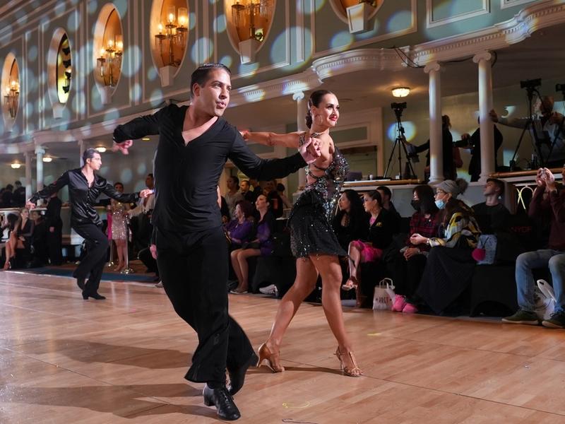 A Latin couple mid-heat on the New York Dance Festival competition floor, spectators and photographers watching from ringside.