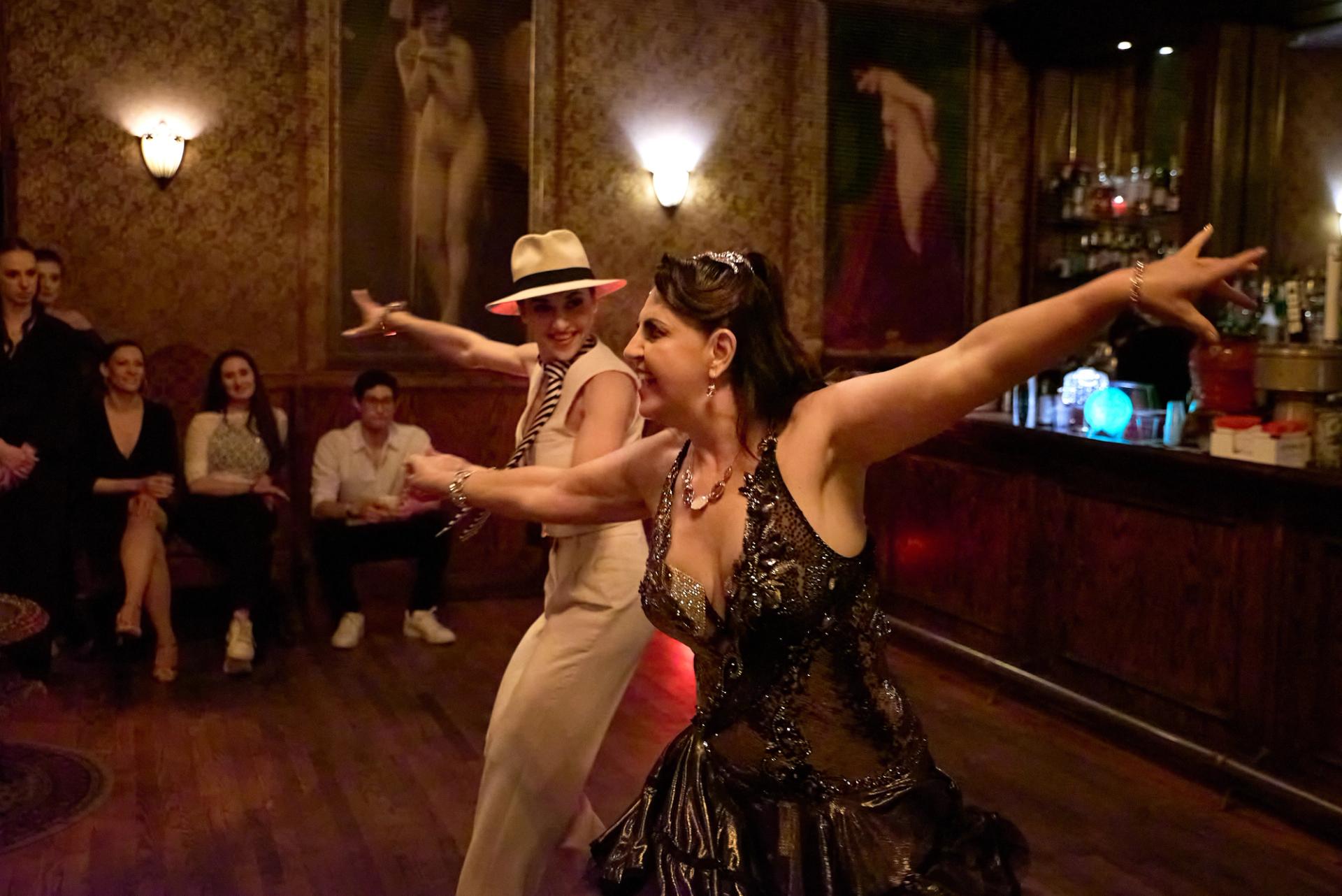 Two dancers arms spread wide mid-cha-cha at the NY Nightlife Showcase — white suit and panama hat beside a sequined gown.