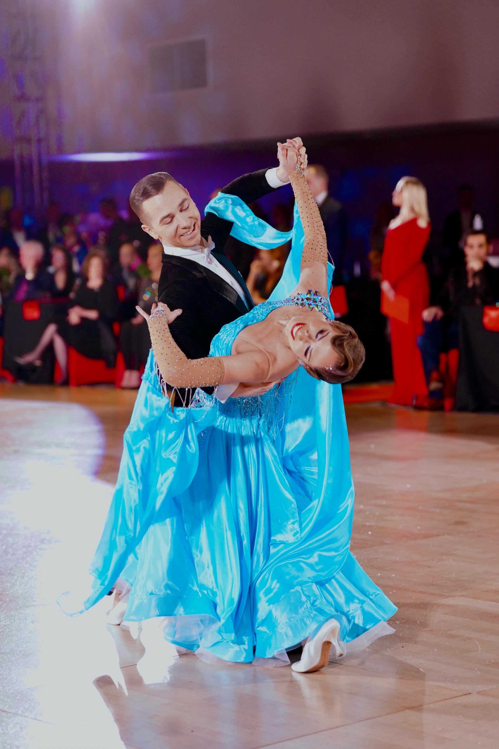 Jaren and Enxhi in an oversway on the competition floor — black tailsuit and a turquoise ballgown mid-swirl.