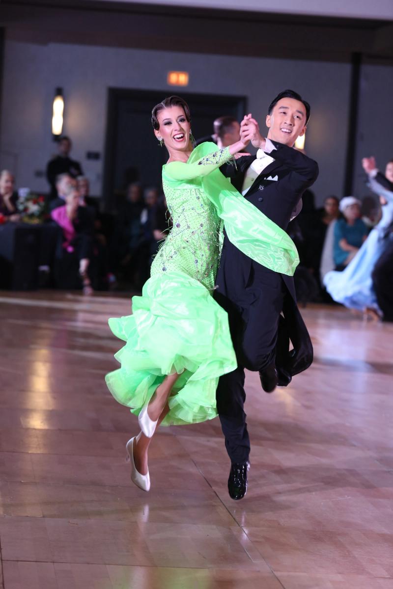 A couple airborne mid-hop on a ballroom floor, the follower in a bright green ruffled gown, the leader in a tailsuit.