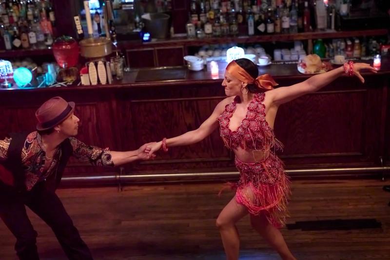 A couple dancing Merengue in front of a lit bar, the follower in a red fringed Latin dress and orange headscarf extending one arm wide while the leader in a fedora and paisley shirt holds her hand.