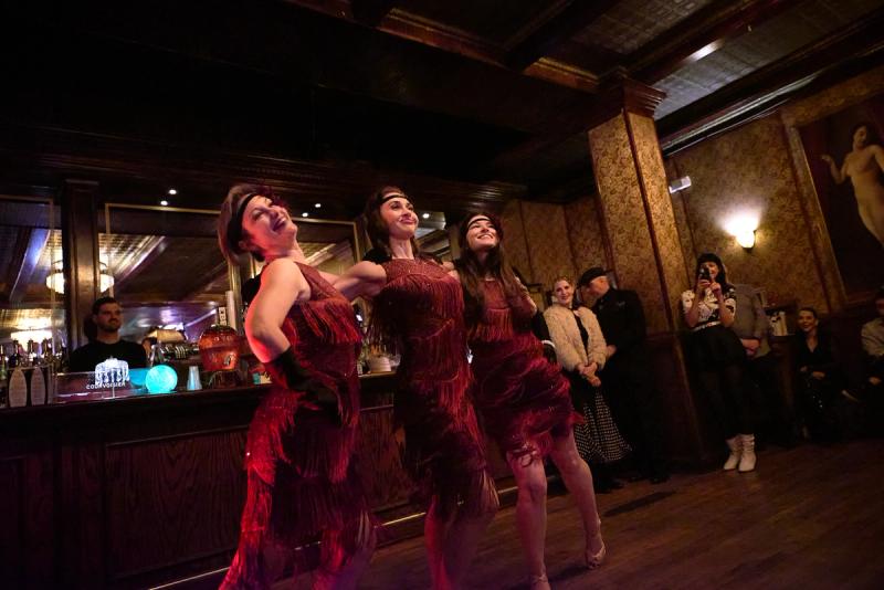 Three dancers in fringed 1920s dresses and headbands performing the Charleston in a wood-panelled speakeasy, with seated guests watching.