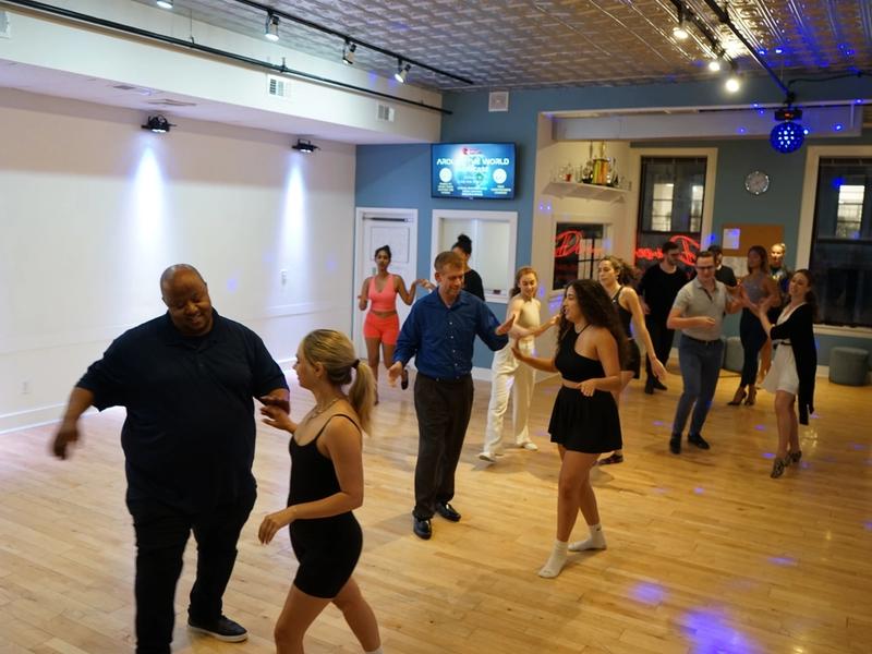 A bright group class at Big Apple Ballroom — students of all ages practicing together on the wooden floor.