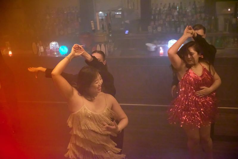 A couple dancing Cha Cha under warm red light at a bar, the follower in a champagne fringed dress with one arm raised in a hand-to-hand hold, a second couple beside them.