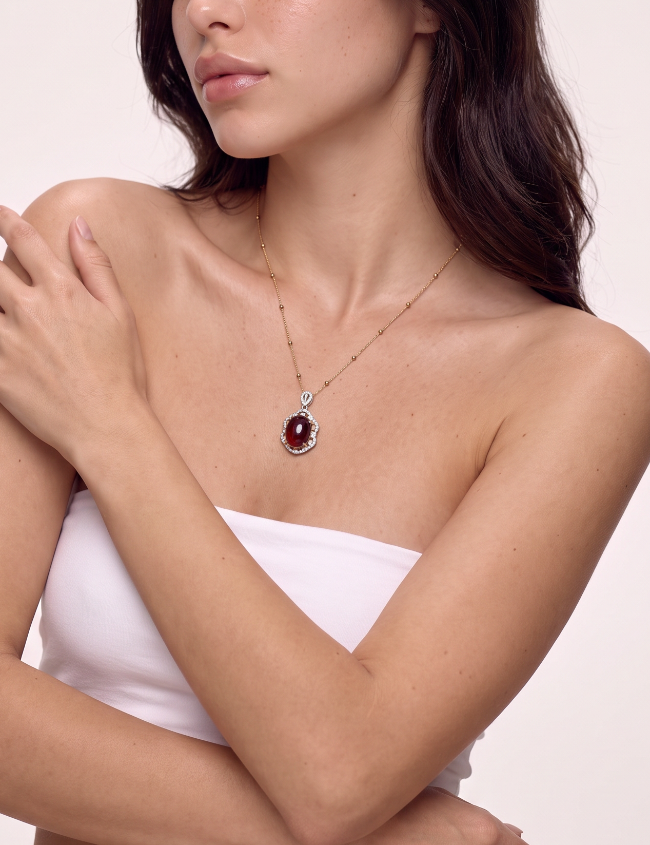 Deep red garnet pendant on a gold chain worn at the neckline, arms folded