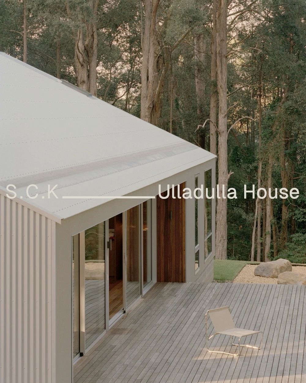 White corrugated house and timber deck with a lounge chair against dense forest.