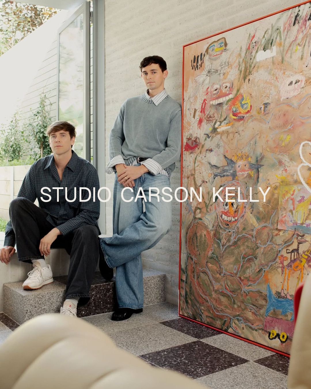 Two people in a sunlit studio beside a large figurative painting in a red frame.