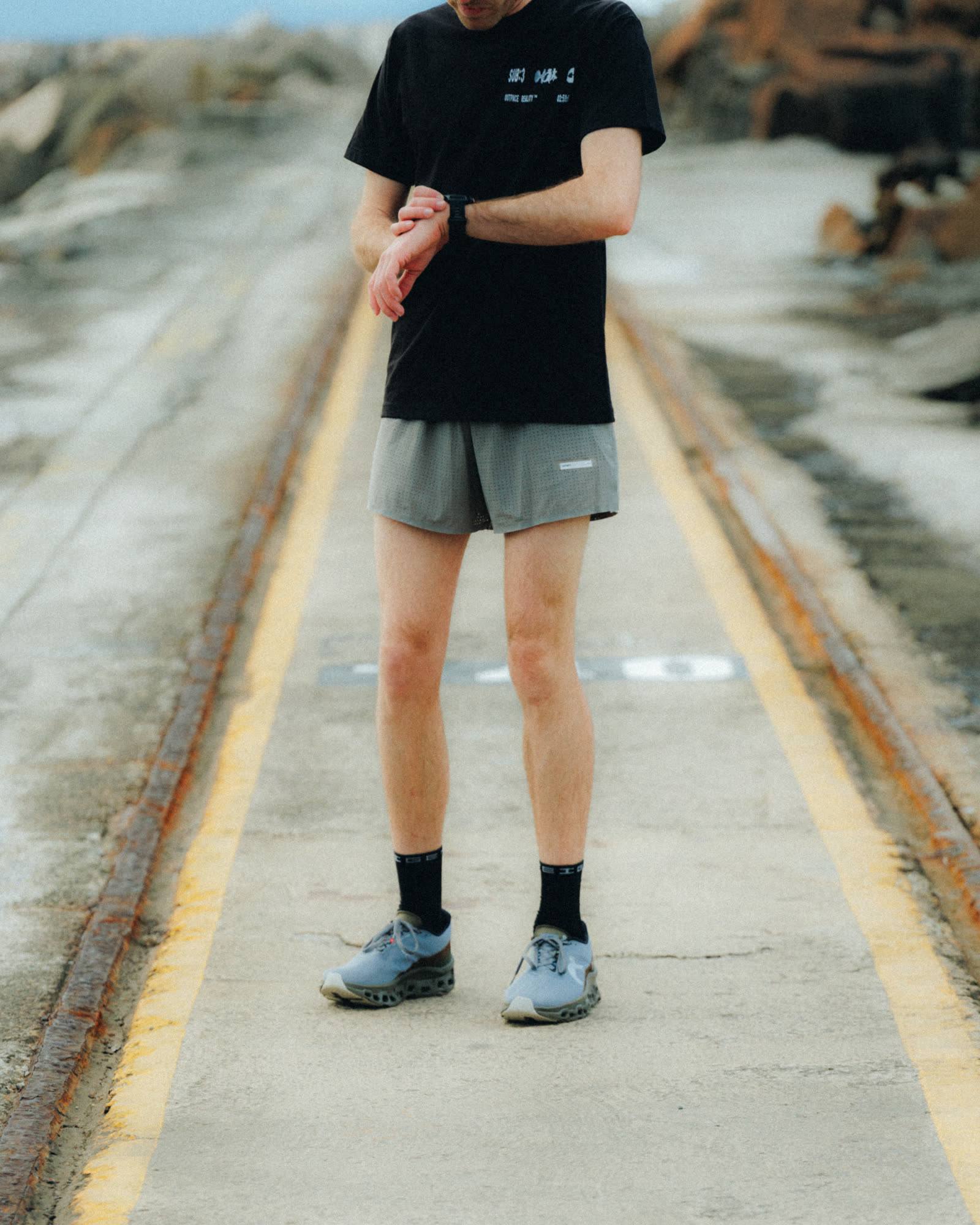 A runner in a black SUB:3 tee on a worn road, checking the watch on their wrist.