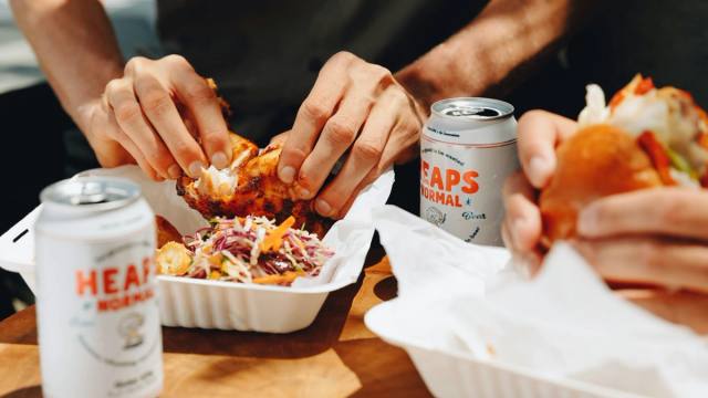 Hands sharing a takeaway meal across a table, two cans of Heaps Normal open beside it.