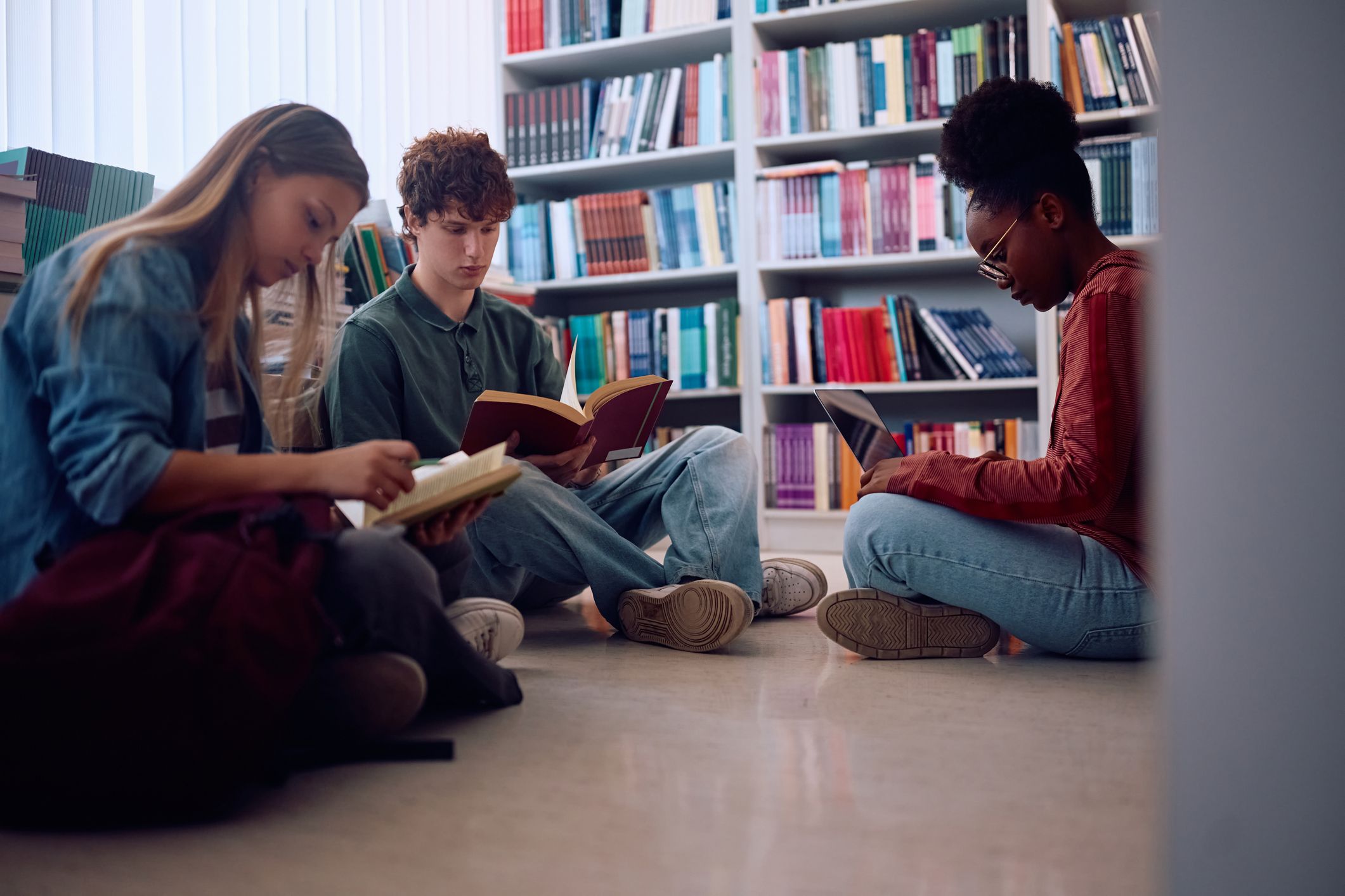 Tre elever på videregående sitter og leser i et skolebibliotek. Bilde.