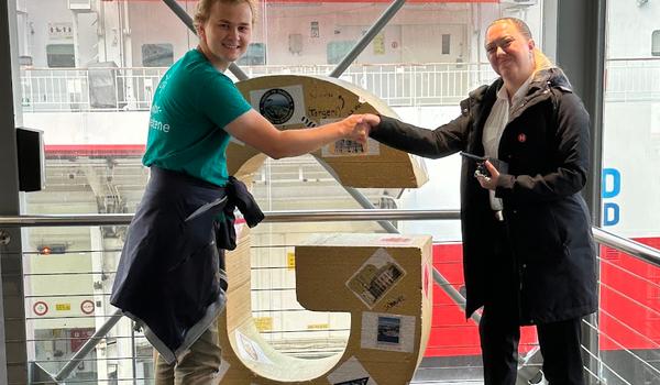 En lektorstudent overrekker en 1,5 meter høy G til en ansatt på Hurtigruten i Bergen.